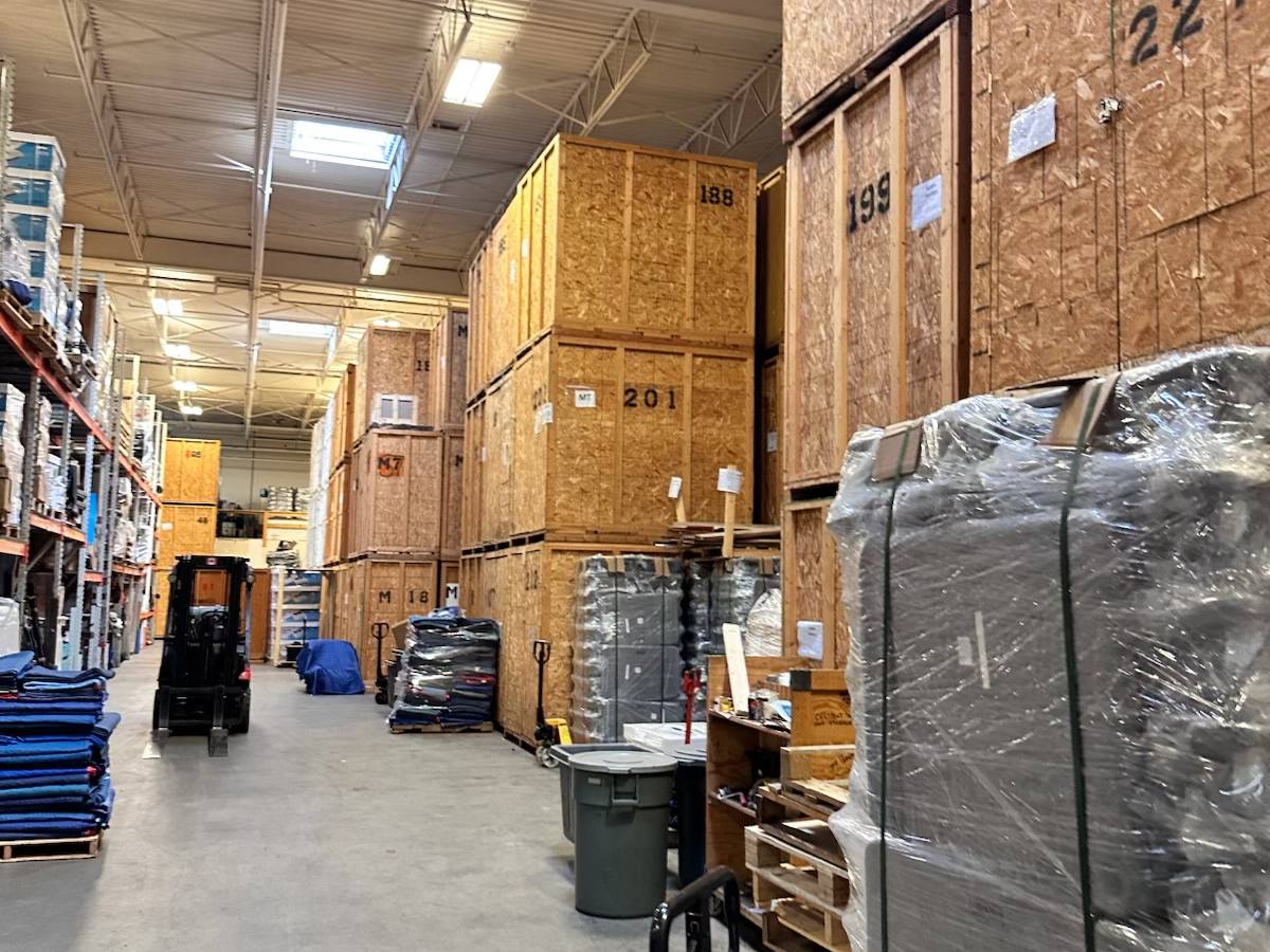 Numbered wooden storage vaults, a forklift and stacks of moving blankets in a warehouse aisle, photographed on one of our jobs in April 2026